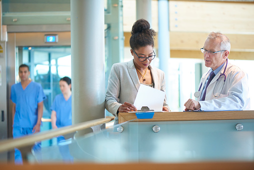 A suited woman and a male doctor wearing a lab coat stand on a stairwell of a modern hospital and discuss some case notes. The omwan could be an administrator or business woman .