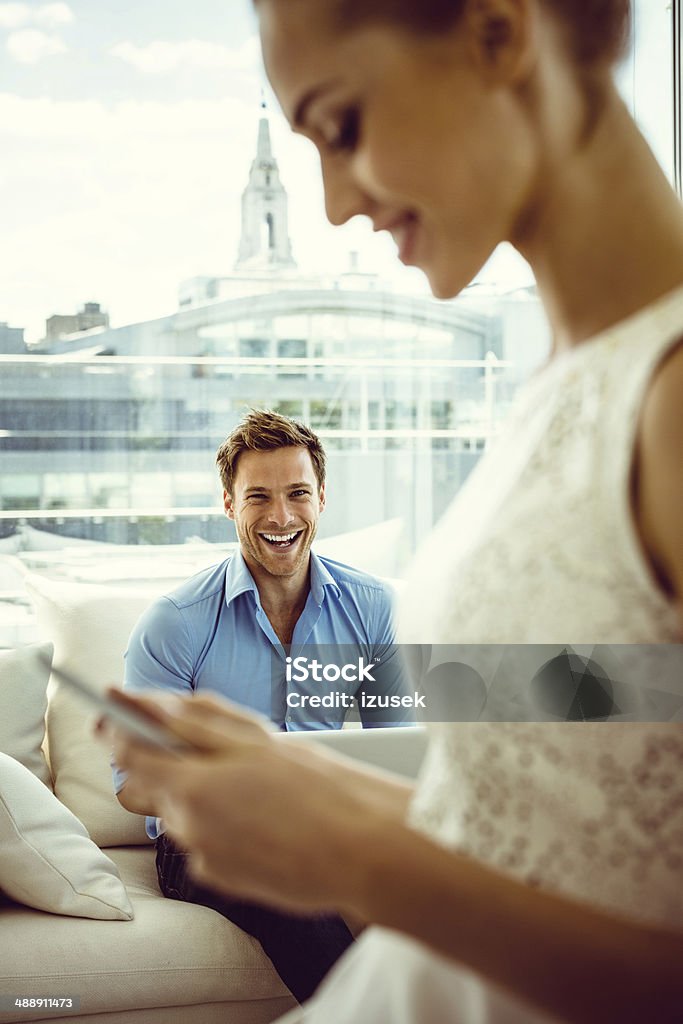 Smiling couple with technologies Focus on young man sitting on sofa in the apartment and using a laptop with the side view of a women texting on the foreground. 25-29 Years Stock Photo Smiling couple with technologies Focus on young man sitting on sofa in the apartment and using a laptop with the side view of a women texting on the foreground. 25-29 Years Stock Photo