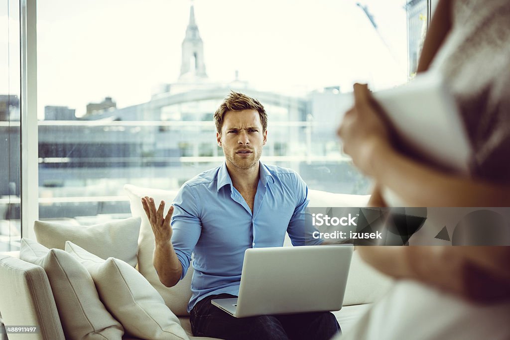 Conflict in family Focus on young man sitting on sofa in the apartment with a laptop on his knees showing off irritation.  Envy Stock Photo