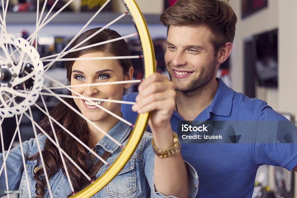 Young woman with wheel Close up of happy young woman holding a bicycle wheel in bike store with her boyfriend or sales clerk standing behind her.  20-24 Years Stock Photo