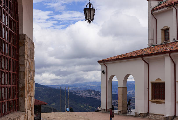 Bogota, Colombia - April 23, 2014:  The view from on top of the Andean peak, Monserrate; below is the capital city of Bogota.  Some visitors sit on the steps of the church and admire the view. Dramatic cloud formations decorate the sky.  A lamp that is suspended from the building on the left seems to almost hand unaided from the sky. To the right is part of the church and a couple of arches that forms one end of the verandah in front of the church on the peak.  Photo taken in the early afternoon.  Horizontal format; copy space.