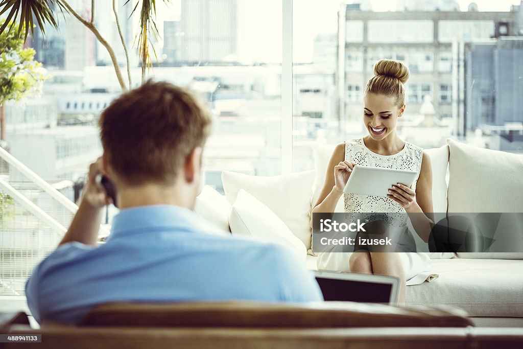 Woman with digital tablet Focus on young woman sitting on sofa at home and using a digital tablet with the back side view of a men talking on phone on the foreground. Digital Tablet Stock Photo