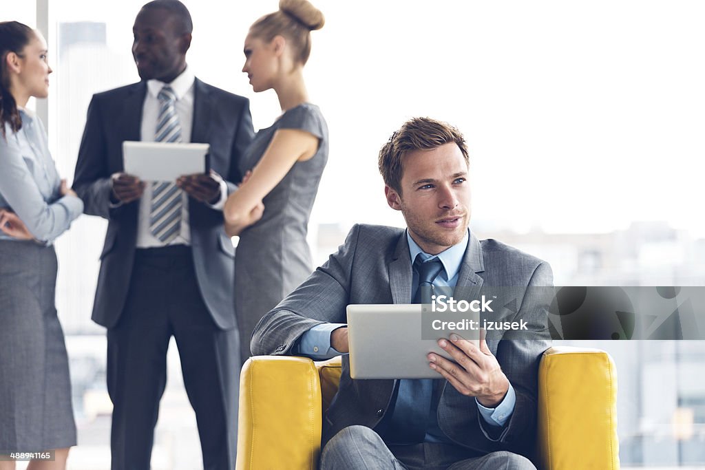 Businessman with digital tablet Focus on the businessman sitting in a chair and using a digital tablet with a group business people talking in the background. 20-24 Years Stock Photo