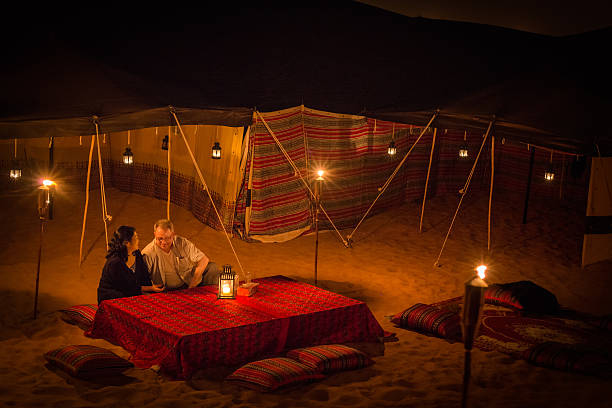 Mature Couple Enjoying Arabian Desert Camping with Lanterns and Torches Dubai iStockalypse.  A mature, multi-ethnic couple enjoy a private, luxury, traditional Arab, Middle Eastern camp in the red desert between Dubai to Sharjah, United Arab Emirates.  The desert camp is lit by candles, lanterns, and torches amid the tents in the sand dunes and is decorated with traditionally patterned pillows, fabrics, carpet and tablecloth. tourist couple candid travel stock pictures, royalty-free photos & images