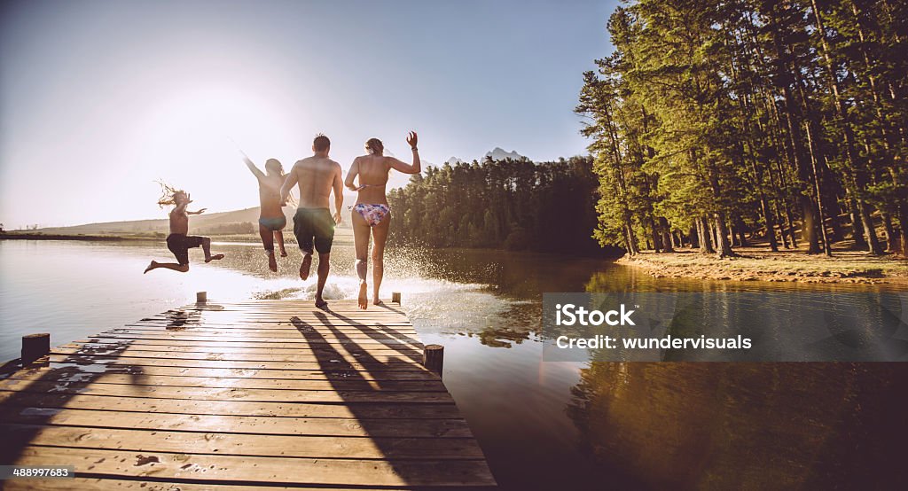 Four people jumping off a jetty into the water A group of people jumping into the water from a jetty or dock.  The dock is made from brown wooden planks. The water is a murky dark color.  The water reflects the trees on the bank, and a warm sun glows on the horizon. Lake Stock Photo