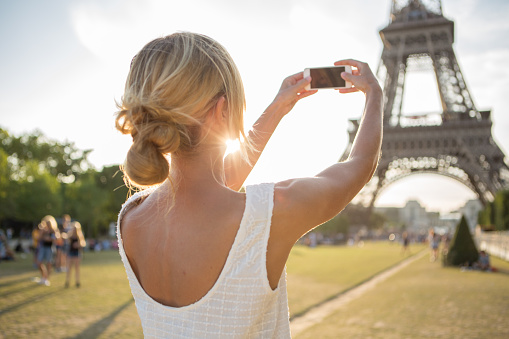 Young cheerful woman near the Eiffel tower  in Paris, France taking a selfie using her mobile phone. Sunset in the capital city.