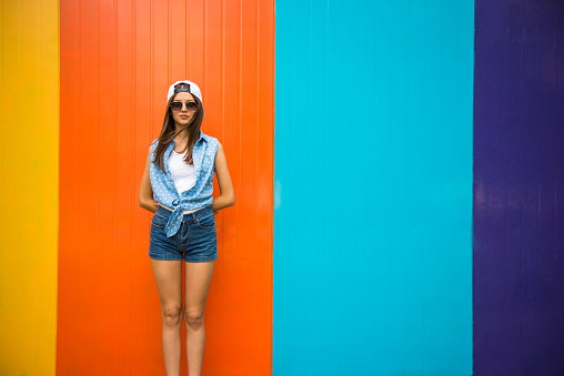 Pretty cool girl in sunglasses and cap standing against the colorful wall.