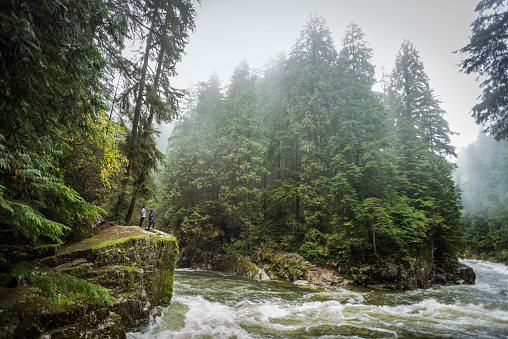 A mature Asian mother and her teenage daughter enjoy the pristine wilderness landscape of a quickly flowing river through a wooded park on a rainy, misty, autumn day from the edge of a small cliff.  Wide angle view of the Capilano River, North Vancouver, Canada.