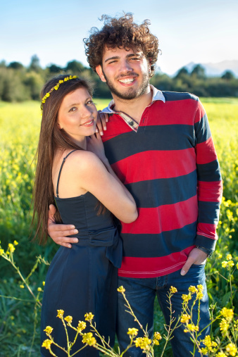 Smiling beautiful romantic couple standing arm in arm in a field of colorful golden yellow rapeseed smiling happily at the camera on a hot summer day.