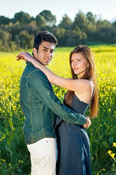 Romantic young couple in an intimate embrace turning to look at the camera as they standing in a colorful golden yellow rapeseed field in summer sun.