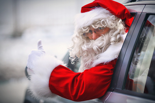 Closeup of modern Santa Claus driving SUV instead of rain deer carriage. He's looking at camera over his shoulder through the rolled down window and showing thumbs up. Blurry snowy background.