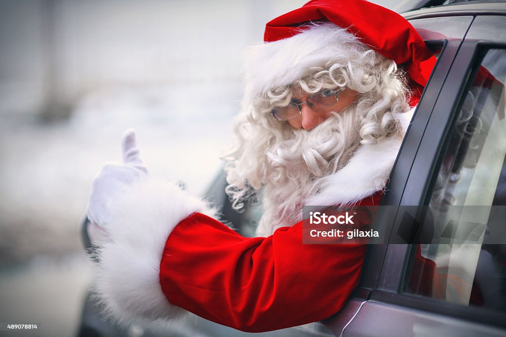 Driving Santa. Closeup of modern Santa Claus driving SUV instead of rain deer carriage. He's looking at camera over his shoulder through the rolled down window and showing thumbs up. Blurry snowy background. Santa Claus Stock Photo