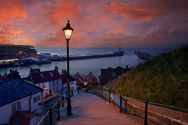 Whitby at dusk - 199 steps leading down from Abbey Whitby is a seaside town, port and civil parish in the Borough of Scarborough and English county of North Yorkshire. Prior to local government reorganizations in the late 1960s it was considered to be part of the North Riding of Yorkshire. Situated on the east coast of Yorkshire at the mouth of the River Esk, Whitby has an established maritime, mineral and tourist heritage. Its East Cliff is home to the ruins of Whitby Abbey, where Caedmon, the earliest recognized English poet, lived. The fishing port emerged during the Middle Ages and developed important herring and whaling fleets, and was (along with the nearby fishing village of Staithes) where Captain Cook learned seamanship. north yorkshire photos stock pictures, royalty-free photos & images