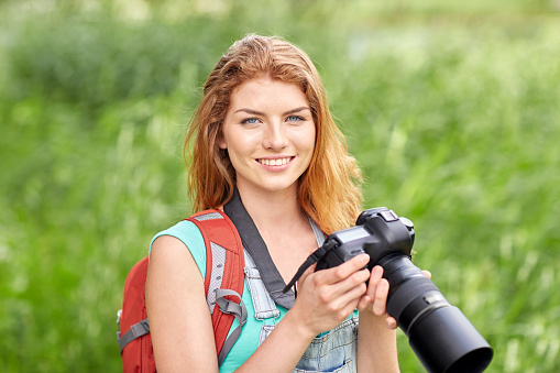 adventure, travel, tourism, hike and people concept - happy young woman with backpack and camera photographing outdoors