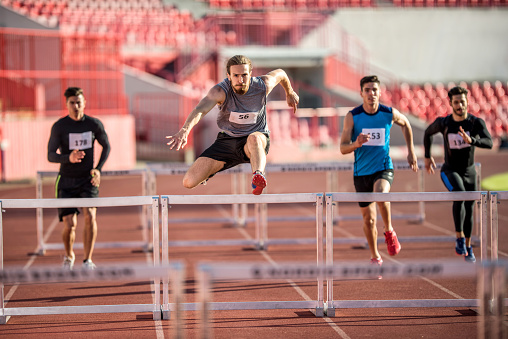Four young man jumping hurdles on a sports race at stadium.