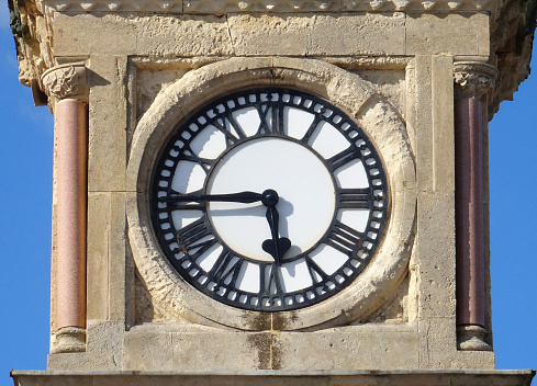 Photo showing a sandstone clock tower with a white clockface, where the numbers around the outside are black Roman numerals.