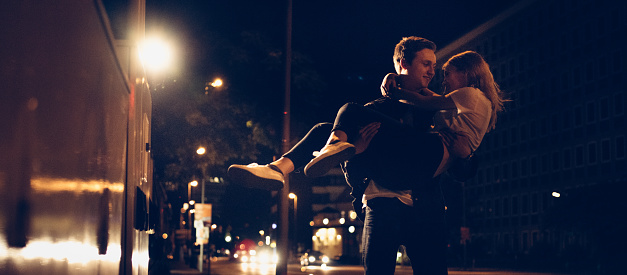 Loving guy carrying his young girlfriend while walking down an urban sidewalk under the city street lights at night