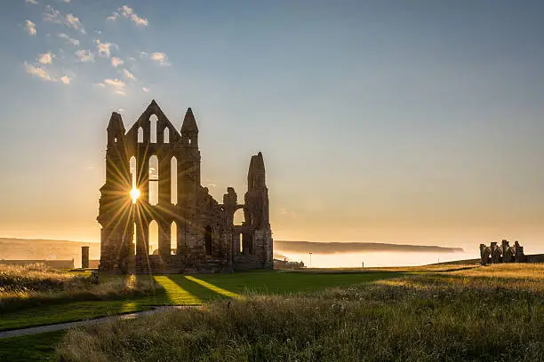The Gothic ruins of Whitby Abbey with the suns rays producing a star.  Sea mist held back by the cliffs at Sandsend in the background