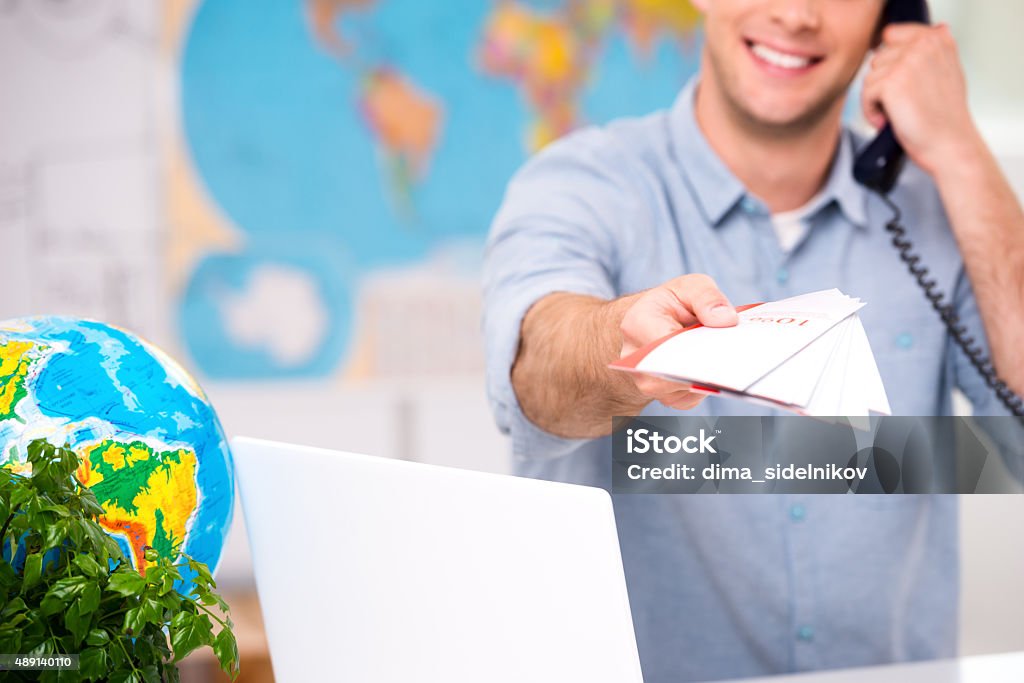 Creative concept for travel agency office Close up photo of male travel agent. Young man talking on phone, smiling, proposing tourist booklet and looking at camera. Travel agency office interior with big world map. Focus on booklet 2015 Stock Photo