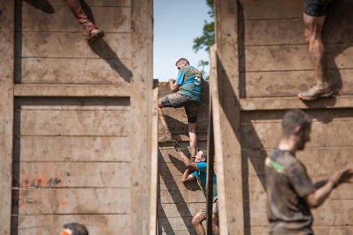 Сirencester, England - August 22, 2015: Man helping a colleague come down a large wooden obstacle as viewed through 2 other fences during his gruelling 12 mile assault course during the ‘Tough Mudder’ challenge in Cirencester park, Gloucestershire. This annual event is a team-based 10-12 mile obstacle course designed to test physical strength endurance.