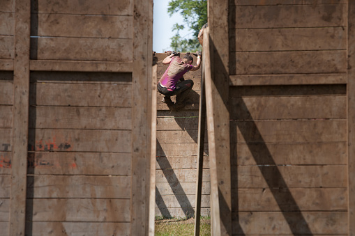 Сirencester, England - August 22, 2015: Woman climbing down a large wooden obstacle as viewed through 2 other fences during her gruelling 12 mile assault course during the ‘Tough Mudder’ challenge in Cirencester park, Gloucestershire. This annual event is a team-based 10-12 mile obstacle course designed to test physical strength endurance.