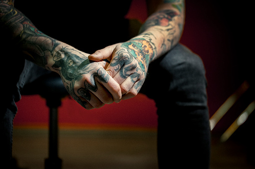 A photo of man's tattooed hands. Close-up of Love and Rich tattoos. Fashionable tattoo artist is sitting with hands clasped at studio.