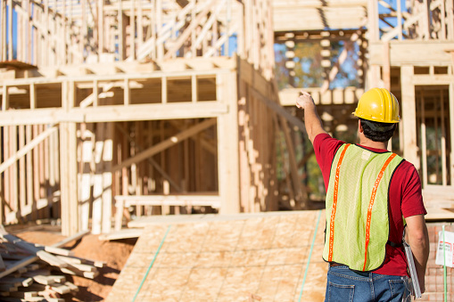 A Latin descent construction worker, supervisor, contractor, inspector, architect, or engineer inspects the work at a construction site.  Framed house, building in background. Lumber.  He is wearing a hard hat and a safety vest and holds a clipboard as he points towards the framed building.  Rear view.
