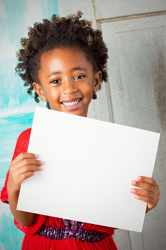 Beautiful elementary age Ethiopian girl with curly hair is smiling and looking at the camera. Child is wearing a red traditional Ethiopian dress. Little girl is holding a blank white sign in front of her.