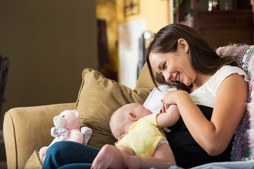 Young adult Caucasian brunette woman is sitting on sofa at home in living room. She is holding her six month old baby daughter while breastfeeding her. Mother is smiling down at daughter while nursing her.