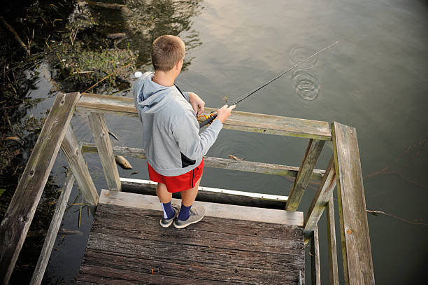 Young teenage man or boy fishing in calm water from end of pier, view looking down.