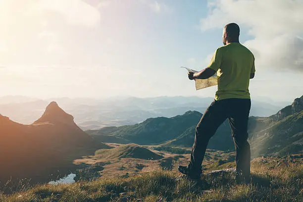 Finding the way. Man with a map standing on top of the mountain, looking ahead.