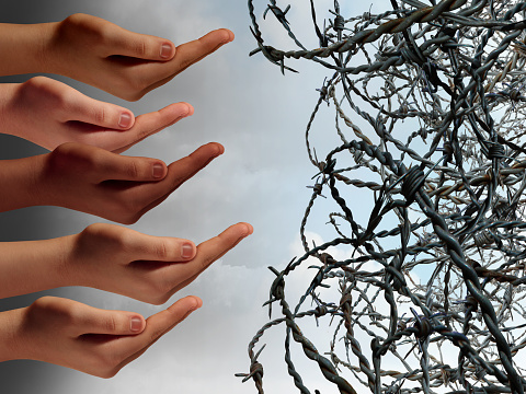 Refugee crisis concept as a group of hands from refugees in distress reaching with open hands asking for help faced with barbed wire fence keeping the suffering people out as a global social issue symbol.