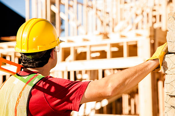 Construction worker busy working at job site. Framed building. Materials. A Latin descent construction worker, supervisor, contractor, inspector, architect, or engineer inspects the work at a construction site.  Framed house, building in background. Pallets of stone to right.  He is wearing a hard hat, gloves, and a safety vest. Rear view. helmet hardhat protective glove safety stock pictures, royalty-free photos & images