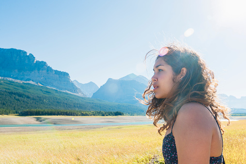 This is a horizontal, color, royalty free stock photograph of a millennial age young woman in her 20s visiting United States travel destination, Glacier National Park in Montana. She stands in the grassy area along Lake Sherburne and looks out over the scenic landscape. The wind blows her hair as the afternoon sun casts a lens flare over the scenic terrain. The cloudless sky is a blue. Many Glacier area mountains fill the background of the rugged landscape. Photographed with a Nikon D800 DSLR camera on a warm autumn day from a side view.