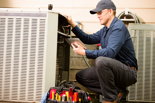 Repairmen works on a home's air conditioner unit outdoors. He is checking the compressor inside the unit using a digital tablet.  He wears a navy blue uniform and his safety glasses.  Tools inside toolbox on ground. 