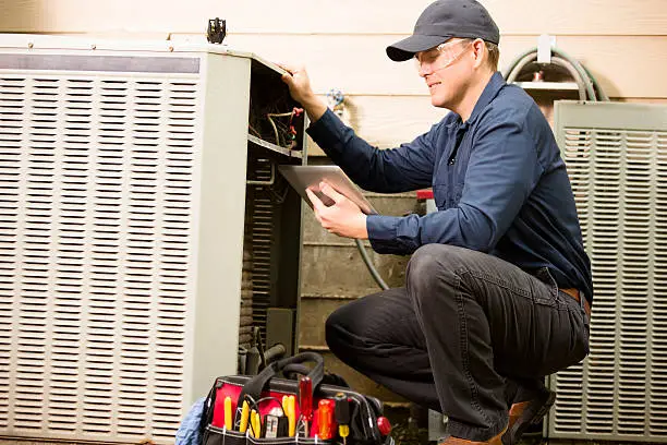 Repairmen works on a home's air conditioner unit outdoors. He is checking the compressor inside the unit using a digital tablet.  He wears a navy blue uniform and his safety glasses.  Tools inside toolbox on ground. 