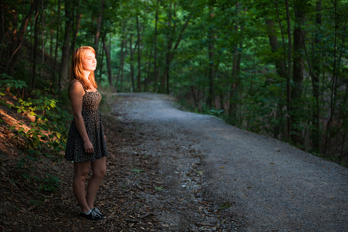 Young woman standing on dark forest path illuminated by evening sunshine