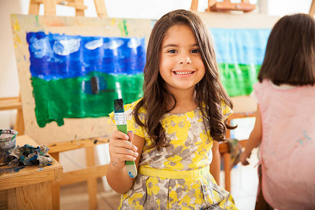 Happy Latin girl in art class Cute Latin girl holding a paintbrush and smiling during art class at school preschool building photos stock pictures, royalty-free photos & images