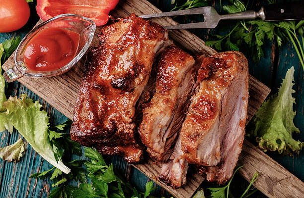 Delicious barbecued ribs seasoned with a spicy basting sauce Delicious barbecued ribs seasoned with a spicy basting sauce and served with chopped fresh vegetables on an old rustic wooden chopping board in a country kitchen. Top view. rib stock pictures, royalty-free photos & images