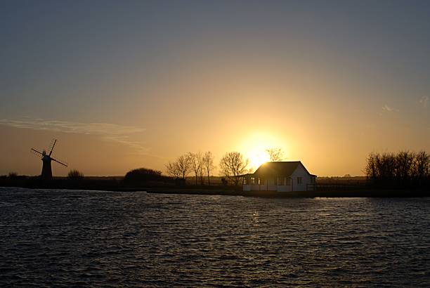Beautiful sunset over the Norfolk Broads with a silhouetted house, windmill and trees.