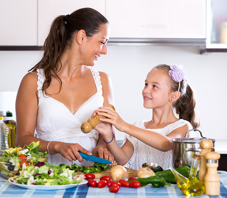 Beautiful little daughter helping mother to prepare stewed vegetables in kitchen