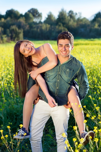 Two attractive laughing young lovers in a field of yellow rapeseed with the man giving his girlfriend a piggy back ride as they enjoy nature together.