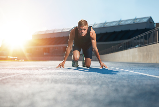 Young man athlete in starting position ready to start a race. Male sprinter ready for a run on racetrack looking at camera with sun flare.