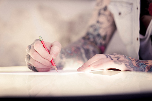 Close-up of male artist's tattooed hands drawing on paper. Professional is holding pencil. He is working in studio.