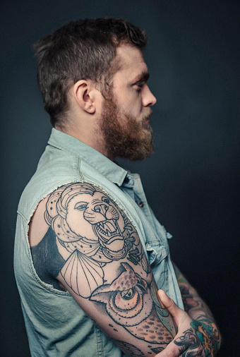 A side view photo of confident fashionable tattooed man. Male is standing against grey background. Hipster is with animal tattoos on arm.