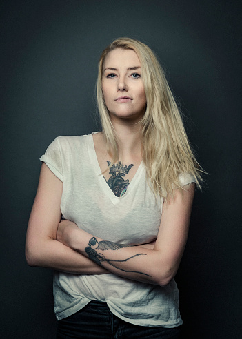 A photo of fashionable young tattooed woman. Portrait of stylish female standing arms crossed. Attractive woman is showing attitude while posing against grey background.