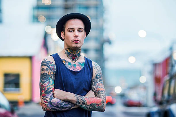 Confident tattooed man standing on city street A photo of confident tattooed man standing arms crossed on city street. Portrait of fashionable male wearing hat and tank top. Hipster is with various colourful tattoos. chest tattoos for men designs stock pictures, royalty-free photos & images
