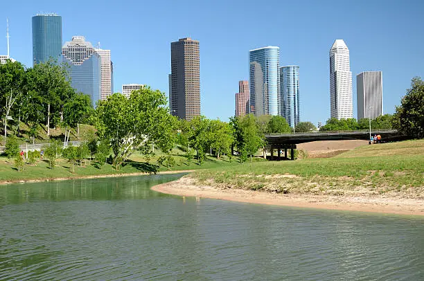 Horizontal, color cityscape of Houston, Texas as viewed across a city park with a waterway in the forground.  At least 10 people exercising by enjoying a bike ride or a walk in the park.