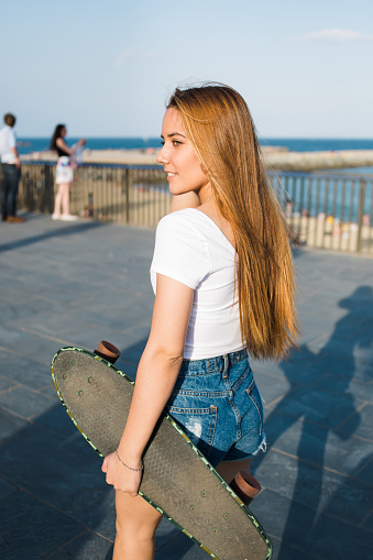 Half length portrait of beautiful woman skateboarder holding her longboard with copy space for your brand while standing on concrete pier in sunny day,