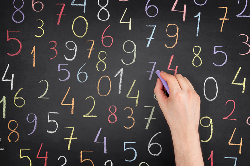 Human hand is writing multi coloured numbers on blackboard.